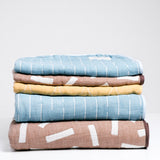 A stack of Blanked Japanese cotton gauze blankets on display at NiMi Projects UK. From top to bottom: a blue small blanket with white line patterning, a small brown version with white rectangles, a small yellow version with white circles, a large blue blanket and a large brown blanket.