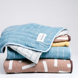 A stack of Blanked Japanese cotton gauze blankets on display at NiMi Projects UK. From top to bottom: a blue small blanket with white line patterning, a small brown version with white rectangles, a small yellow version with white circles, a large blue blanket and a large brown blanket.