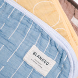 A closeup of three cotton Blanked Japanese gauze blankets fanned out on display at NiMi Projects. From left to right: A pale blue blanket with stitched white lines, a yellow blanket with stitched white circles and a brown blanket with stitched white rectangles.