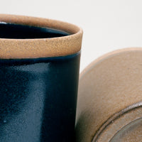 A close up of two NiMi Projects' Sakuzan Japanese ceramic Straight Mugs, showing the unglazed lip of a navy blue mug on the left and the base of a beige-brown mug on the right. 