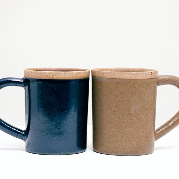 A pair of Japanese ceramic Mino-yaki Sakuzan Straight Mugs, facing each other. One in navy blue (left) and the other in beige brown (right). Each mug has an unglazed lip showing the natural color and texture of the clay, and large rounded handles. Available at NiMi Projects UK.