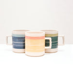 Three Japanese porcelain Mino-yaki Kanna mugs, designed by ceramicist Sone Yoji, each with bands of two-tone colour textured with tiny indented flecks (a pottery technique called tokibanna). Colours from left to right — navy blue with a slimmer band of pale blue, yellow with a orange and dark green with lime green. On display at NiMi Projects UK.