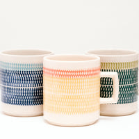 Three Japanese porcelain Mino-yaki Kanna mugs, designed by ceramicist Sone Yoji, each with bands of two-tone colour textured with tiny indented flecks (a pottery technique called tokibanna). Colours from left to right — navy blue with a slimmer band of pale blue, yellow with a orange and dark green with lime green. On display at NiMi Projects UK.