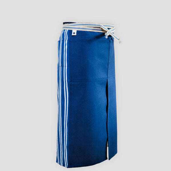 A long Sanpu Sanyo apron in navy blue, as worn, featuring a sailcloth canvas front panel with two pockets and striped pocketed side panels of tenugui cotton. In place of apron strings is a wrap-around sanada himo woven waist cord. Made in Japan and available at NiMi Projects UK.