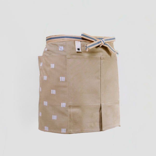A short beige Sanpu Sanyo apron, as worn, with graphic patterned tenugui cotton pocketed side panels, a sailcloth canvas double-pocketed front and sanada himo woven waist cord. Made in Japan and available at NiMi Projects UK