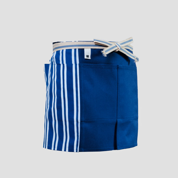A short Sanpu Sanyo kitchen or gardening apron in navy blue, with sailcloth canvas front panel sporting two pockets, striped side tenugui cotton side panels, and a sanada himo wrap-around  waist cord. Made in Japan and available at NiMi Projects UK.