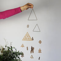 Japanese designer Sukima's wooden Forest mobile, held up to show its size, featuring woodland motifs of a triangular forest, six trees, a deer, a bird, a snowman, a star, a Christmas stocking and two brass triangles.