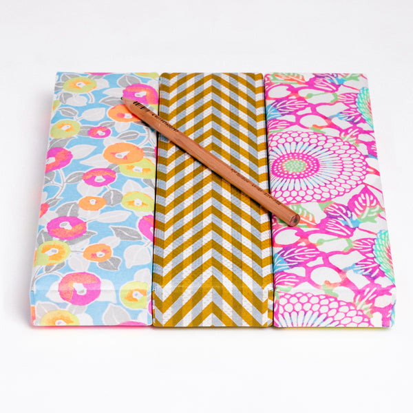 Three Japanese Yuzen Washi Pen boxes on show at NiMi Projects UK, made with traditional chiyogami washi paper that has been screen-printed with colourful patterns. Boxes from left to right — a peony patterned box in neon pink, yellow and orange, a herringbone patterned box in yellow and gray, and a chrysanthemum patterned box in neon pink and blue.