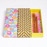 Three Japanese Yuzen Washi Pen boxes, made with traditional chiyogami washi paper that has been screen-printed with colourful patterns. Boxes from left to right — a peony patterned box in neon pink, yellow and orange, a herringbone patterned box in yellow and gray, and an open yellow box with a chrysanthemum pattern in the bottom in neon pink and blue. Available at NiMi Projects UK.
