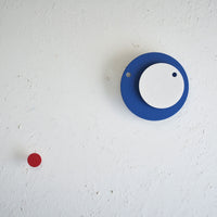 A Fukunaga Paper Clock of two circles, a blue one with a white one on top, displayed on a white wall with a small red decorative dot to its lower left.