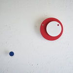 A Fukunaga Paper Clock of two circles, a white one on top of a blue one, on display on a white wall, with a decorative blue dot place below left.
