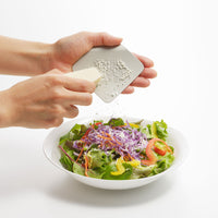 A silver Japanese irogami kitchen grater being held and used with a block of parmesan cheese over a bowl of salad.