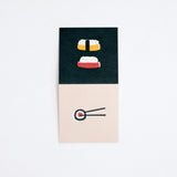 A Japanese Motif Mini Card, laid flat to show both sides, with one side showing  picture of two pieces of nigiri sushi on a black background, and a pair of chopsticks holding a norimaki sushi roll on a cream background on the other side. Printed in Japan and available at NiMi Projects.