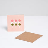  A square Japanese Motif Mini Card, featuring an illustration of three sticks of hanami dango (pink, white and green mochi balls) on a pink background, with a brown envelope. Printed in Japan and available at NiMi Projects UK.