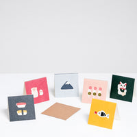 A collection of six square Japanese Motif Mini Cards, each featuring an illustration of a Japanese symbol, at NiMi Projects UK. Clockwise from the left: A black card with two nigiri sushi, a red card with two white monkeys, a blue card with Mount Fuji, a pink card with three sticks of hanami mochi dango, a green card with a white manekineko lucky cat and a yellow card with a fish-shaped bottle of soy sauce.