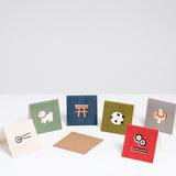  A collection of six square Japanese Motif Mini Cards, each featuring an illustration of a Japanese symbol, at NiMi Projects UK. Clockwise from the left: a white card wth a norimaki and pair of chopsticks, a green card with a white dog, a blue card with a red torii gate, a green card with a mochi cake, a grey card with a yellow uchide no kozuchi lucky mallet and a red card with two norimaki sushi rolls and a pair of chopsticks.