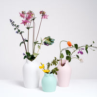Three figure-like Ceramic Japan x Nendo Dress Up vases, each with various summer flowers. From left to right — a large vase in white with a wing tip collar as the vase neck, a small vase in pale green with a Peter Pan collar, and a medium vase in pink with a dropped scalloped neckline. All against the white background of the NiMi Projects UK store.