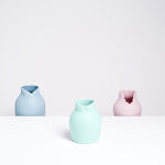 A Ceramic Japan x Nendo pastel green porcelain small Dress Up vase, with a Peter Pan collar at its neckline, so that it appears like a rotund figure. Behind it are two other Dress Up vases on a lower shelf - a blue-gray version with a wing-tip collar on the left and a pink version with a scalloped dress collar on the right. Made in Japan and displayed at NiMi Projects UK.