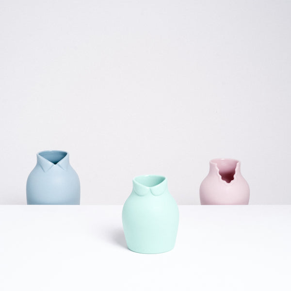 A Ceramic Japan x Nendo pastel green porcelain small Dress Up vase, with a Peter Pan collar at its neckline, so that it appears like a rotund figure. Behind it are two other Dress Up vases on a lower shelf - a blue-gray version with a wing-tip collar on the left and a pink version with a scalloped dress collar on the right. Made in Japan and displayed at NiMi Projects UK.