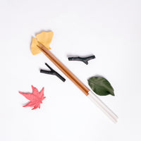 Five Japanese porcelain chopstick rests in the shape of leaves and branches — a yellow ginkgo leaf, pink maple leaf, green cherry-tree leaf and two brown branches. Here displayed with a pair of wooden chopsticks at NiMi Projects UK. 