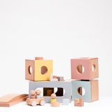 The Dou Toy I'm Home set of building blocks, showing four box blocks with round windows, which can be used as rooms to a doll house.  Other parts include three wooden dolls, a small cart car, L-shaped blocks, mini cylinder and cube blocks, and a curved roof block. All on display at NiMi Projects UK.