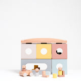 NiMi Projects' Dou Toy I'm Home building block doll house, featuring four square box blocks with round windows in pastel colours, a long block with an oblong window, a curved roof block, two L-shaped blocks, several mini cylinder and cube blocks, three dolls and a cart-like car. 