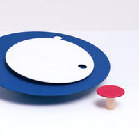 Close up of a  Fukunaga Paper Clock of two circles, a small white one indicating minutes on top of a blue one indicating hours, and an extra decorative small red dot, laid out out on a white table at NiMi Projects UK.