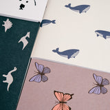 A collection of Japanese notebook covers featuring different motifs. Clockwise from top left — polar bears on white, whales on ivory white, poodles on pink, butterflies on grey and dinosaurs on green.
