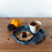 An autumnal display on a wood table of two Mino-ware Japanese porcelain arabesque-edged plates — a small yellow one on top of a large blue one, displayed with a rustic Japanese tea cup. Items available at NiMi Projects UK
