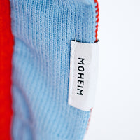 A closeup of the edge of a NiMi Projects' Moheim Silhouette knitted vase cover, showing a sky blue frilled edge with the Moheim label, and orange center. 