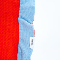 A closeup of the edge of a NiMi Projects' Moheim Silhouette knitted vase cover, showing a sky blue frilled edge with the Moheim label, and orange center. 