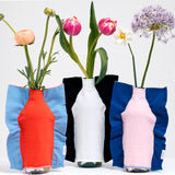 Three NiMi Projects' Moheim Silhouette fine-knitted vase covers, each  stretched over a plastic bottle holding different flowers. From left to right: An orange Silhouette cover with a sky blue background, a white Silhouette cover with navy background and a pink silhouette cover with a blue background.