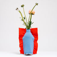 A Moheim Silhouette jersey knit vase cover available at NiMi Projects UK, featuring a sky blue silhouette of a vase on an orange background and stretched over a bottle holding pink peonies.