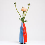 A side view of a Moheim Silhouette jersey knit vase cover available at NiMi Projects UK, featuring an orange silhouette of a vase on a sky blue background and stretched over a bottle holding pink peonies.
