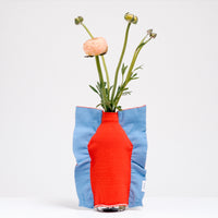 A Moheim Silhouette jersey knit vase cover available at NiMi Projects UK, featuring an orange silhouette of a vase on a sky blue background and stretched over a bottle holding pink peonies.