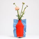 A Moheim Silhouette jersey knit vase cover available at NiMi Projects UK, featuring an orange silhouette of a vase on a sky blue background and stretched over a bottle holding pink peonies.