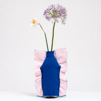 A Moheim Silhouette jersey knit vase cover available at NiMi Projects UK, featuring a blue silhouette of a vase on a pink background and stretched over a bottle holding a purple flower.
