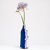 A side view of a  Moheim Silhouette jersey knit vase cover available at NiMi Projects UK, featuring a pink silhouette of a vase on a blue background and stretched over a bottle holding a purple flower.