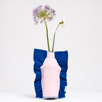 A Moheim Silhouette jersey knit vase cover available at NiMi Projects UK, featuring a pink silhouette of a vase on a blue background and stretched over a bottle holding a purple flower.