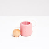 An NiMi Projects pink Ototama Cup and Ball game. This toy is a Japanese kendama-like game featuring a cup and a natural colored ball that’s attached to its center by a white cord.