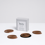 A grey cardboard box of  Trunk Design's Daily Uzumaki outdoor insect repellent spirals, with four of its brown incense coils arranged in front, one on a black heatproof mat. Made in Japan on Awaji Island and available at NiMi  Projects UK.
