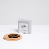 A Trunk Design Daily Uzumaki brown insect repellent incense coil displayed on a heatproof mat in a Trunk Design wooden Incense holder, with a grey box of coils behind. Made in Awaji, Japan, and on sale at NiMi Projects UK.