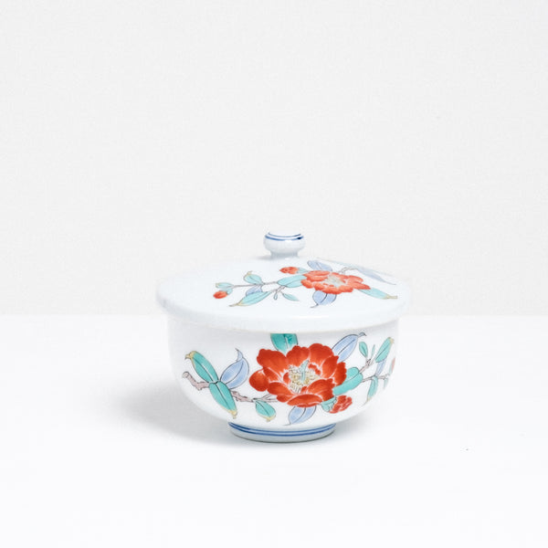 Side view of a white vintage Japanese porcelain lidded tea cup, featuring a design of hand painted red peonies and blue and green leaves.