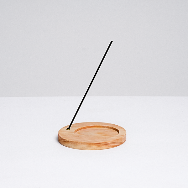 A circular wooden incense stand, with a circle indent for a heatproof mat for paper incense, and a side hole holding a stick of traditional Japanese incense, made in Japan by Trunk Design and available at NiMi Projects 